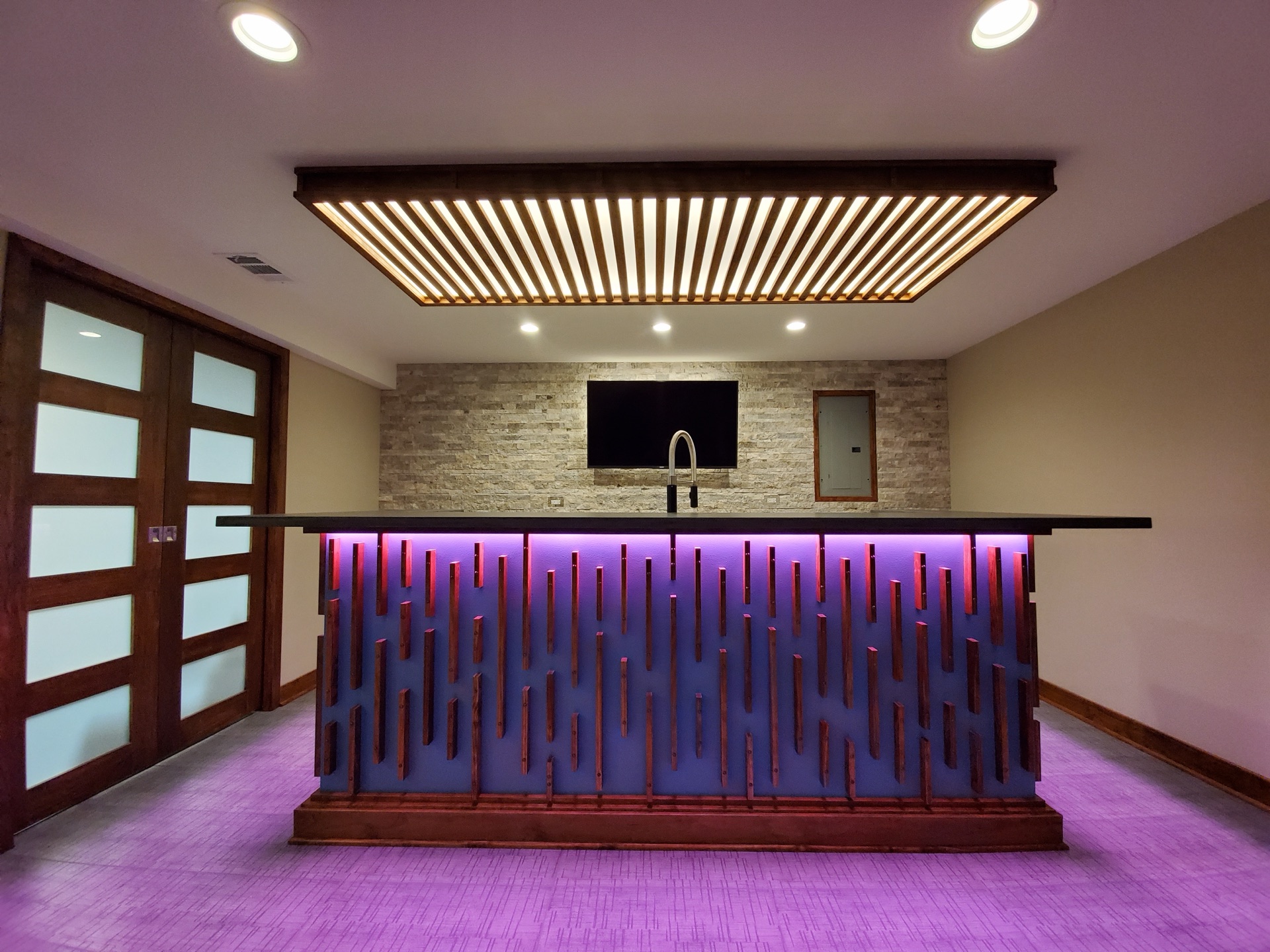 Detail of custom slat-front basement bar with under-counter lighting and wood-grid ceiling feature by Alpha Development Group