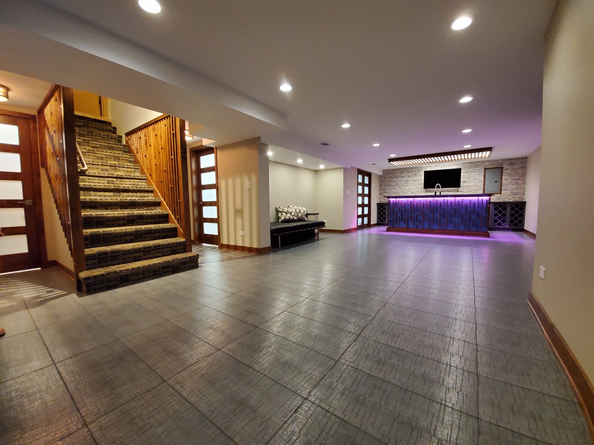 Wide view of custom finished basement: hardwood staircase, mood-lit bar, hex slate flooring, mounted TV, padded bench seating
