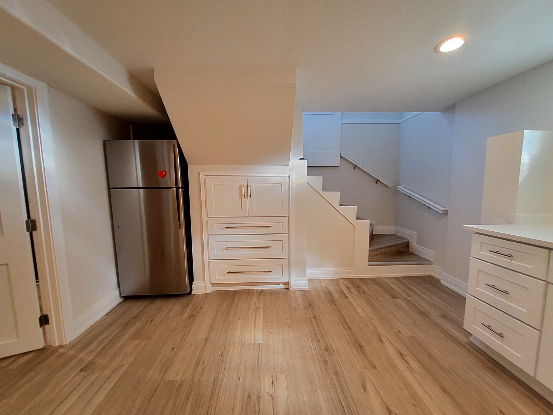 Custom basement kitchenette by Alpha Development Group: full-height stainless fridge, white cabinetry, hardwood floors, recessed lighting